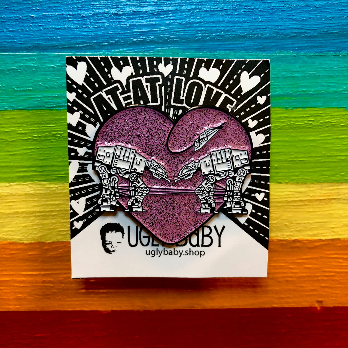 Heart shaped enamel pin called Love AT-AT First Sight with a pink glitter background. Shown against a rainbow background.