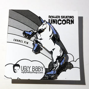 A roller skating unicorn enamel pin mounted on its original white backing card. The backing card features a black and white illustration of a unicorn on a rainbow with the text 'ROLLER SKATING UNICORN' and the brand 'UGLY BABY' at the bottom.