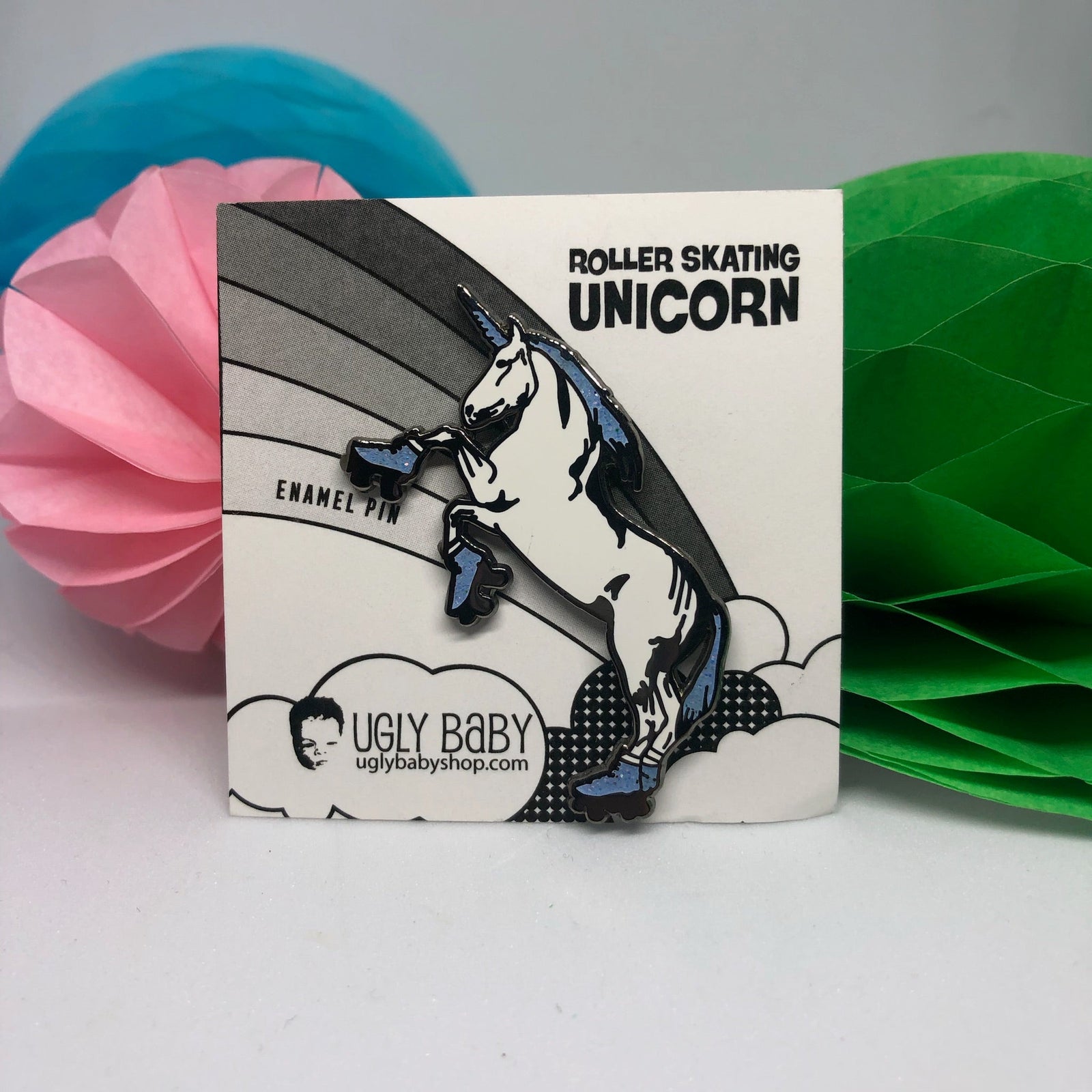 A roller skating unicorn enamel pin mounted on its original white backing card. The backing card features a black and white illustration of a unicorn on a rainbow with the text 'ROLLER SKATING UNICORN' and the brand 'UGLY BABY' at the bottom.