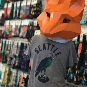 A heather gray shirt that says SEATTLE in block letters. Below there is a seahawk wearing a helmet and standing on a football. It is shown on a mannequin wearing an orange fox mask made of paper. In the background there is a wall of socks blurred. 