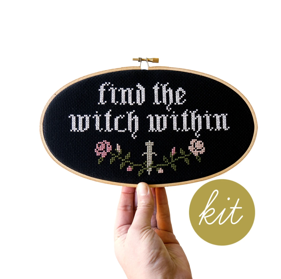 A finished example of the Find the Witch Within Cross Stitch Kit by Junebug and Darlin. It is being held up by a human hand against a white background. 