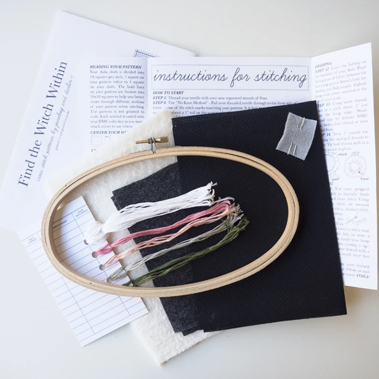 An overhead shot of the supplies included in the Find the Witch Within cross stitch kit. You get an embroidery hoop, needles, aida fabric, embroidery floss, felt, a pattern and hand illustrated instructions. 