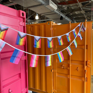 Colorful rainbow flags hanging between two shipping containers, one pink and one orange.