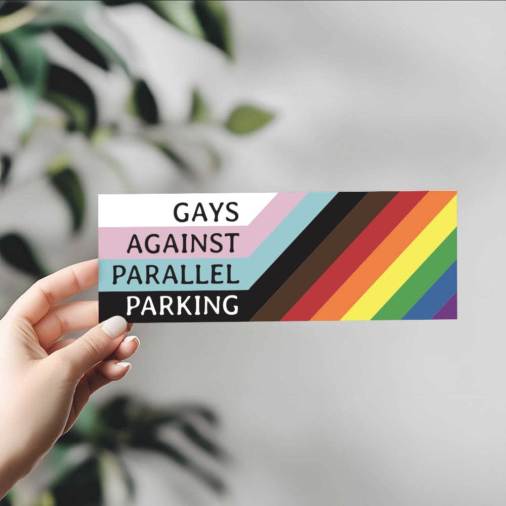 Hand holding a rainbow-colored sticker with text 'GAYS AGAINST PARALLEL PARKING' against a blurred green leafy background.