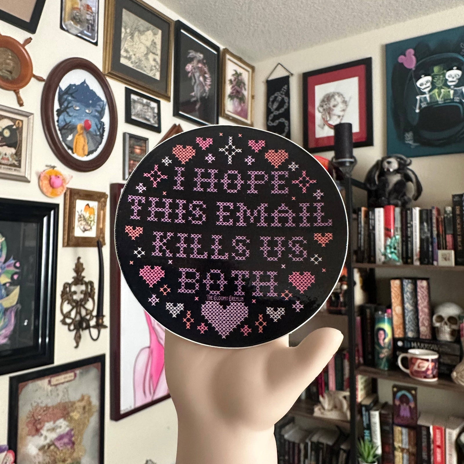 A hand holds a round cross-stitch sticker with the text 'I HOPE THIS EMAIL KILLS US BOTH' in pink and purple thread on a black background, set against a cozy, cluttered room with framed art and bookshelves.