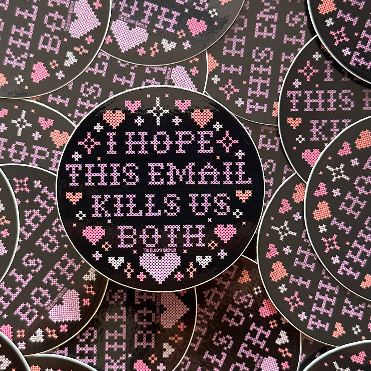 A circular cross-stitch style sticker with a black background and pink and white stitched text reading 'I HOPE THIS EMAIL KILLS US BOTH,' surrounded by repeated matching circular designs in the background.