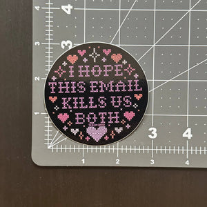 A round black sticker with pink and white cross stitch text reading 'I HOPE THIS EMAIL KILLS US BOTH' surrounded by small hearts, placed on a gray cutting mat over a dark brown surface.