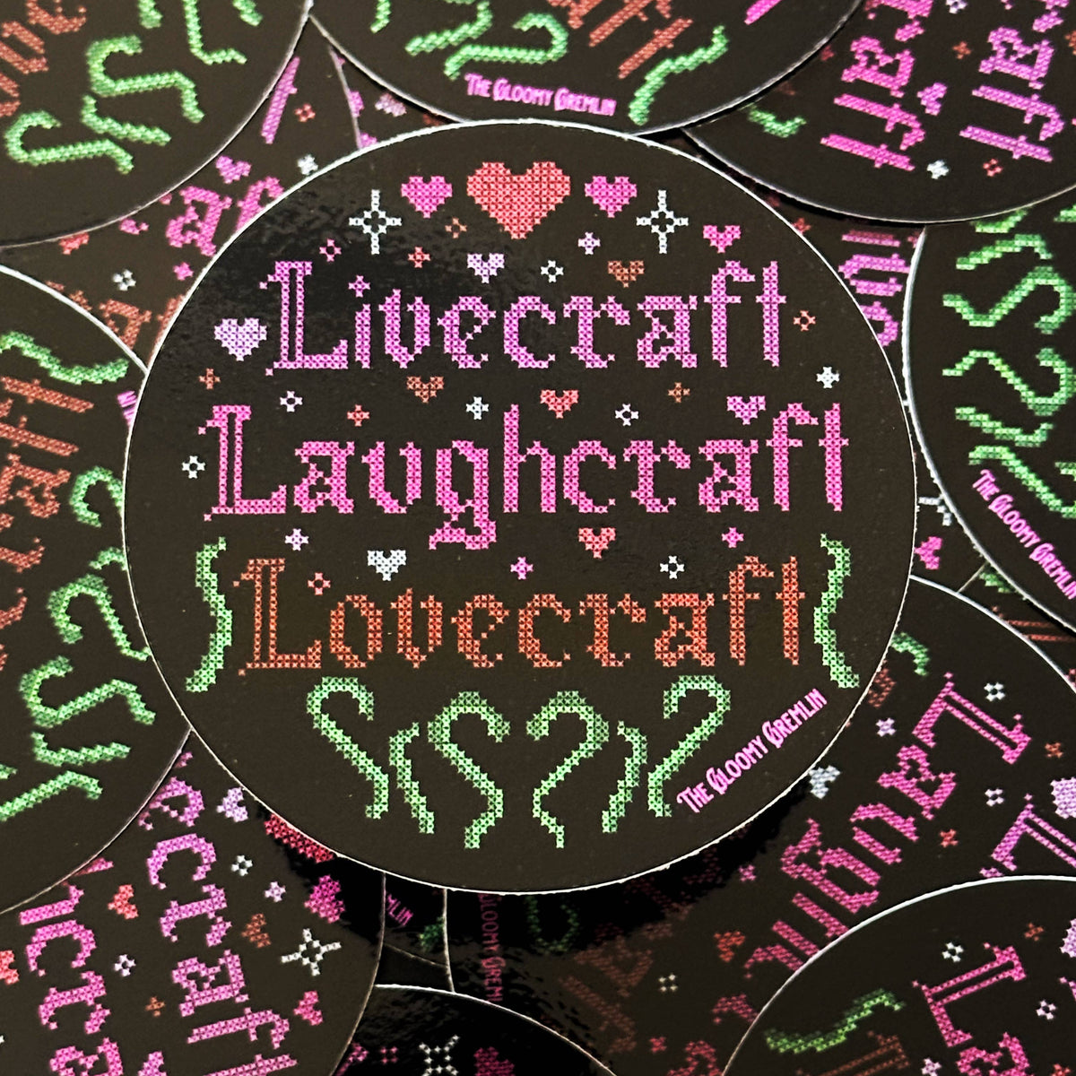 A circular sticker featuring a cross-stitch style design with the words 'Livecraft Laughcraft Lovecraft' in pink, green, and orange on a black background, surrounded by small hearts and decorative motifs. Multiple identical round pieces are arranged overlapping in a flat-lay pattern.