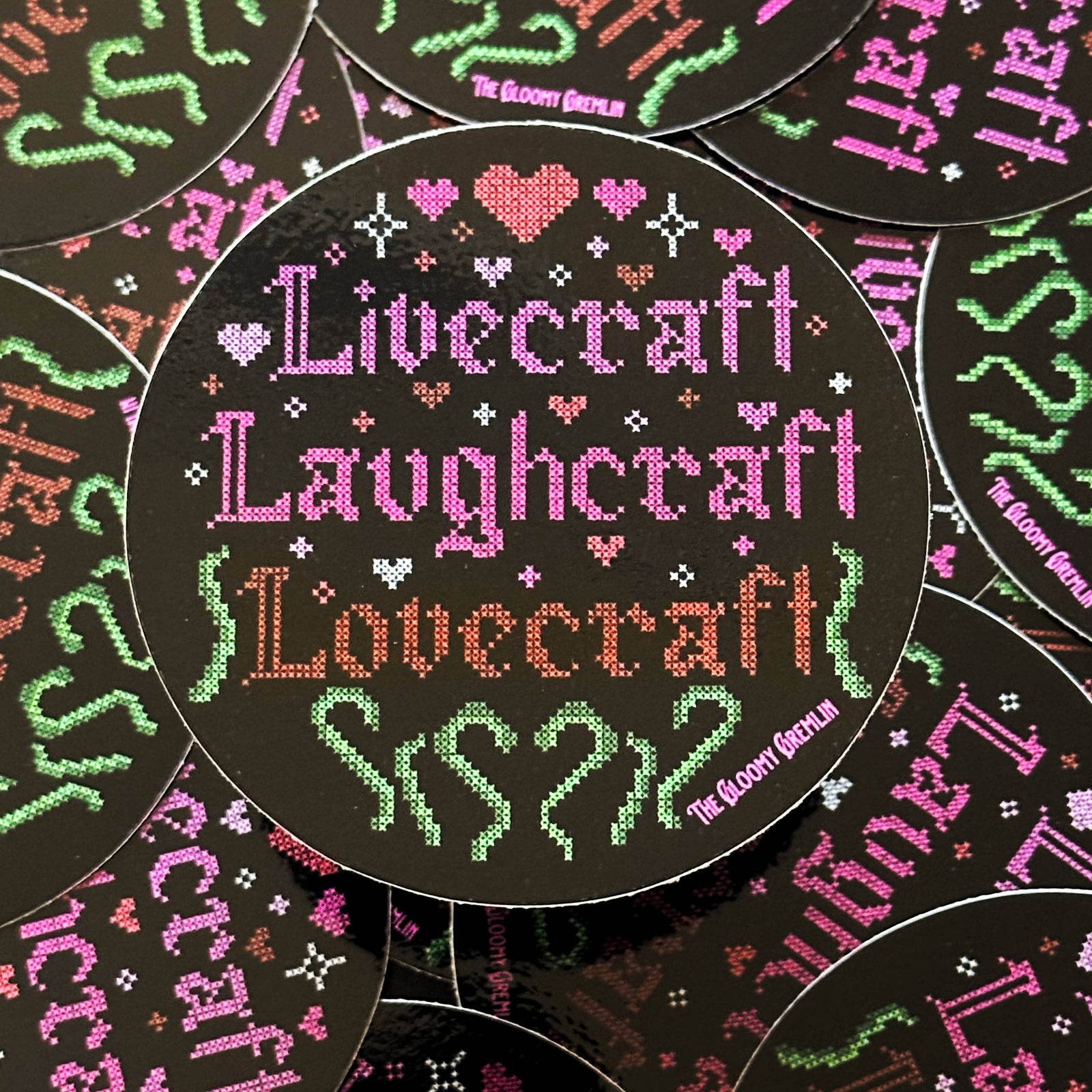 A circular sticker featuring a cross-stitch style design with the words 'Livecraft Laughcraft Lovecraft' in pink, green, and orange on a black background, surrounded by small hearts and decorative motifs. Multiple identical round pieces are arranged overlapping in a flat-lay pattern.