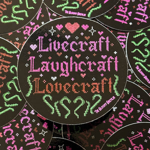 A circular sticker featuring a cross-stitch style design with the words 'Livecraft Laughcraft Lovecraft' in pink, green, and orange on a black background, surrounded by small hearts and decorative motifs. Multiple identical round pieces are arranged overlapping in a flat-lay pattern.