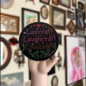 A hand holds a round black sticker featuring the words 'Livecraft Laughcraft Lovecraft'