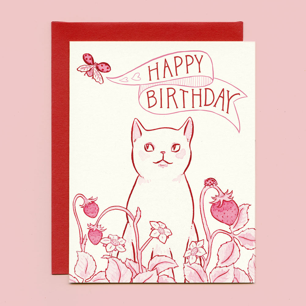 A card that says "Happy Birthday" on a banner and features a cat sitting in a patch of strawberries watching a ladybug flying by. 