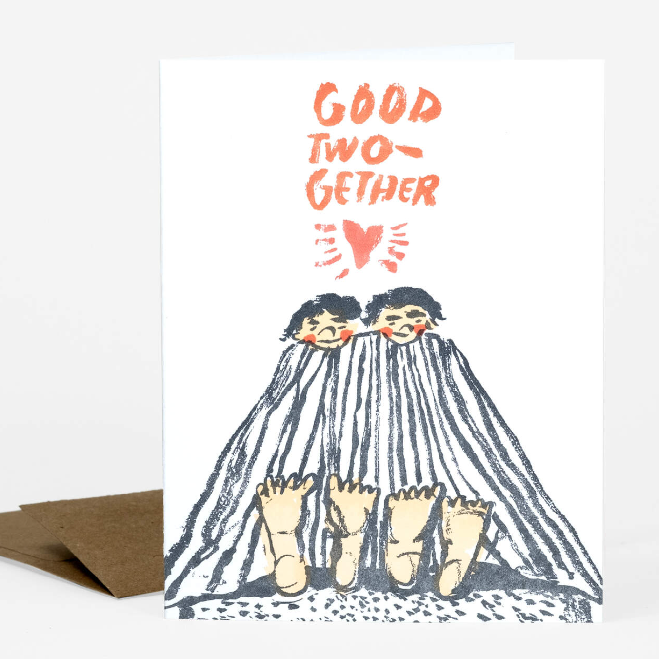 A letterpress greeting card with two dark haired people on the front. They are laying under a blanket and staring at their bare feet poking out the end It says "Good Two-gether"