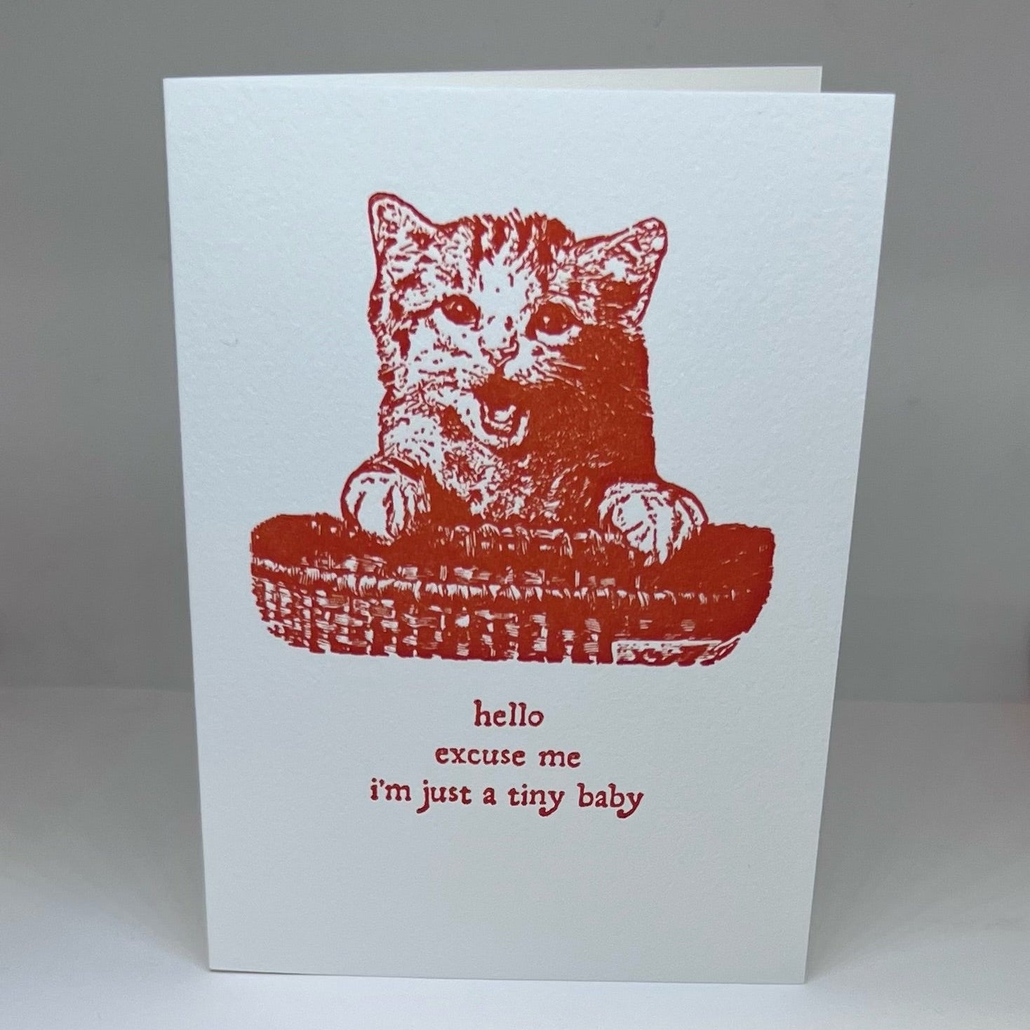White letterpressed greeting card with red cat design and text that says "hello. excuse me. i'm just a tiny baby." on a white background