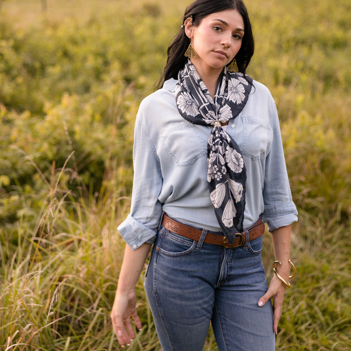 Person wearing a blue button down shirt and jeans modeling a floral patterned bandana against a grassy background. 