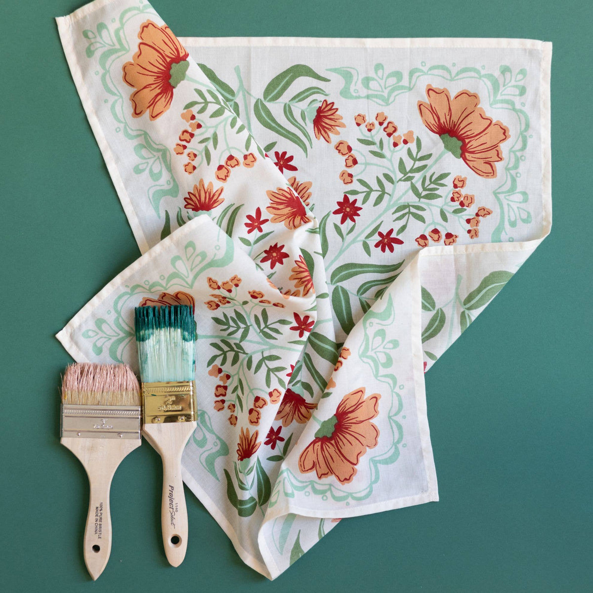 White bandana with an orange and green floral pattern with two paint brushes against a teal background. 