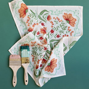White bandana with an orange and green floral pattern with two paint brushes against a teal background. 