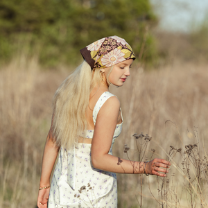 Blonde woman wearing a white dress modeling a floral patterned bandana as a head scarf standing in a field. 