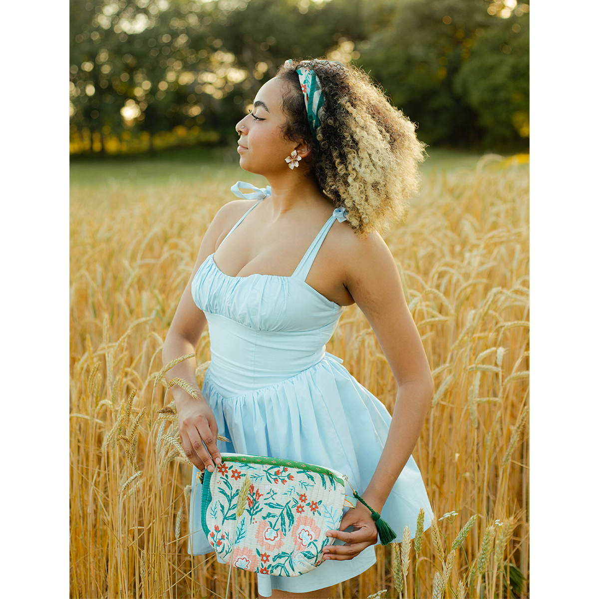A woman wearing a light blue dress and standing in a field, holding a floral patterned pouch and wearing a floral patterned bandana in her hair as a headband. 