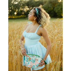 A woman wearing a light blue dress and standing in a field, holding a floral patterned pouch and wearing a floral patterned bandana in her hair as a headband. 