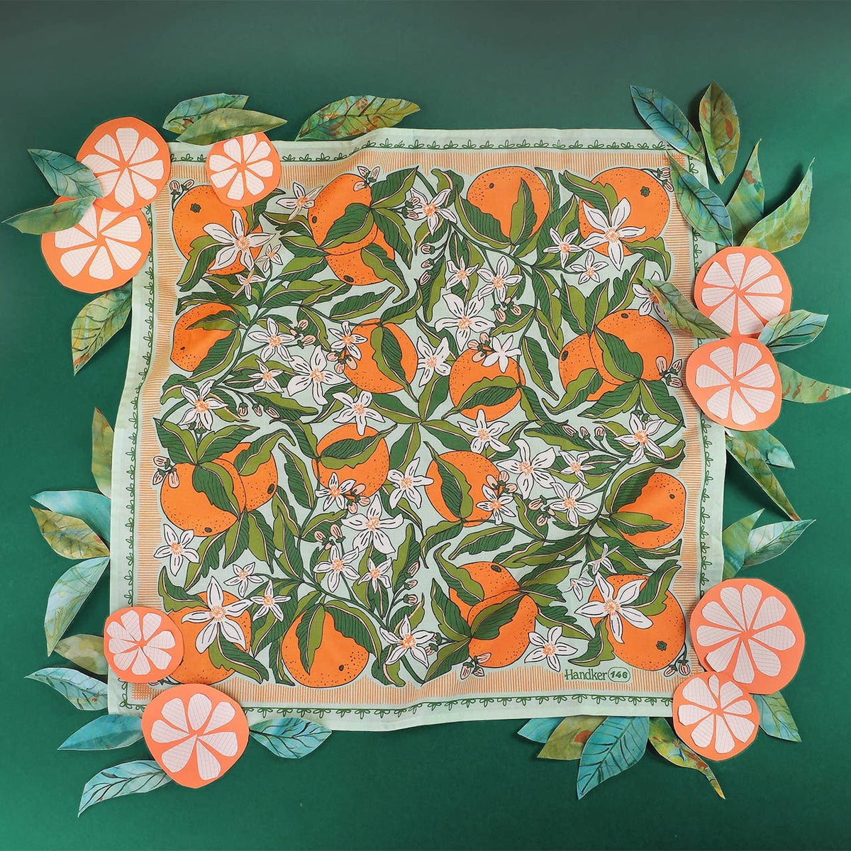 Bandana with an orange and orange blossom pattern laid out on a green background, surrounded by paper cutouts of leaves and orange slices. 