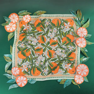 Bandana with an orange and orange blossom pattern laid out on a green background, surrounded by paper cutouts of leaves and orange slices. 