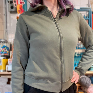 A person wearing an olive green zip-up hoodie with a hood and black pants, standing in a store setting with shelves and colorful items in the background.