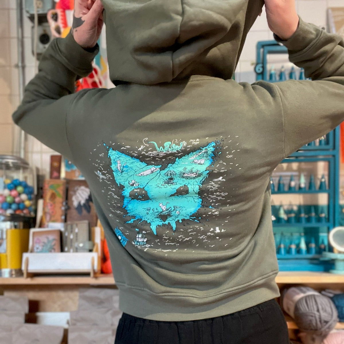A person wearing an olive green hoodie with a large blue and white treasure map in the shape of a cat head graphic on the back, standing in a shop with shelves of yarn and other items in the background.