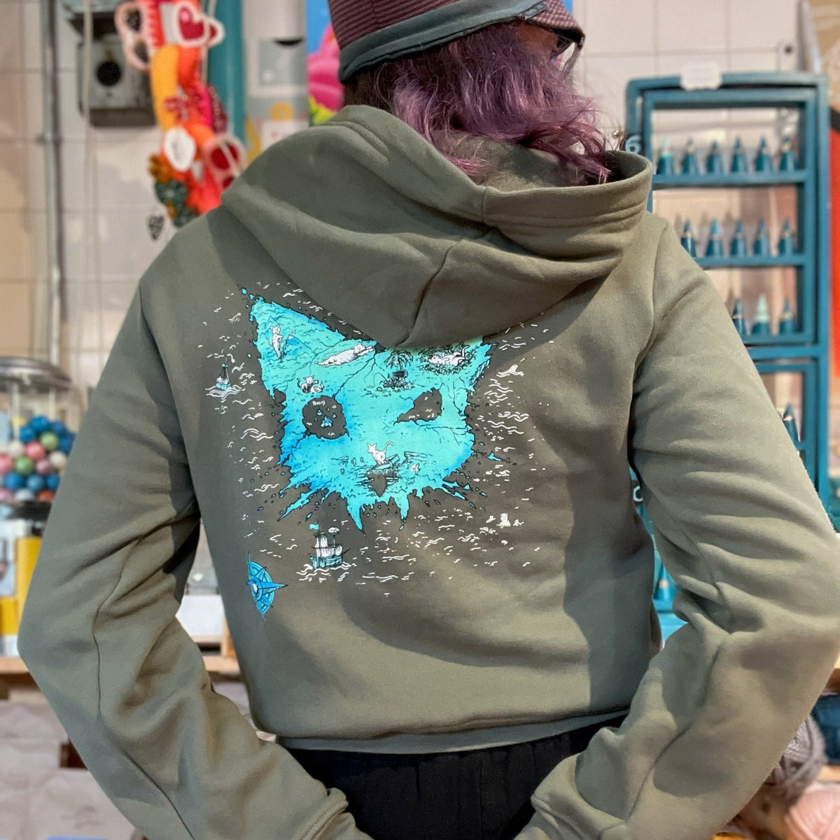A person shown from behind wearing an olive green cropped hoodie with a large turquoise and white treasure map in the shape of a cat head graphic print on the back, standing in a store.