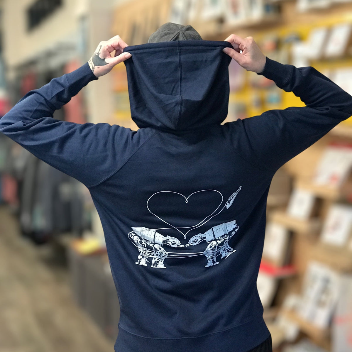 Person wearing a navy blue hoodie with the Love AT-AT First Sight design on the back in an indoor setting.