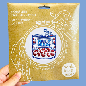 A hand holding a kraft paper package for a complete embroidery kit. The package features white line art and text, including 'COMPLETE EMBROIDERY KIT' and 'FULL O' BEANS', with a circular logo for 'hook, line & tinker'.