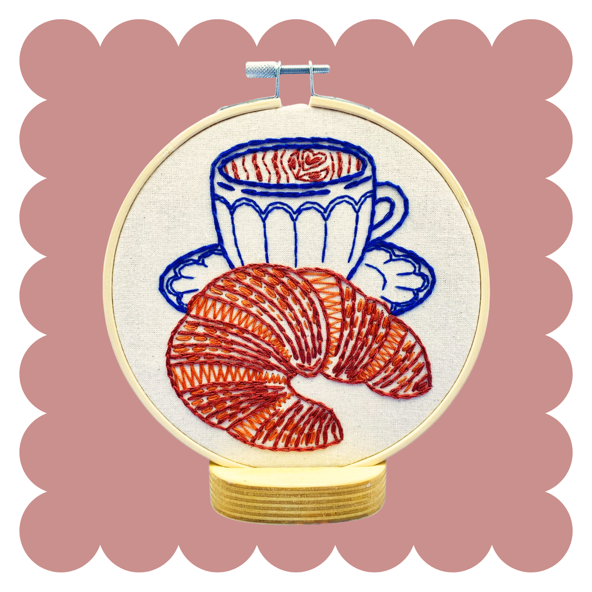A circular embroidery hoop displaying a stitched design of a blue coffee cup and saucer with a croissant on an off-white fabric background, shown on a stand against a dusty pink scalloped backdrop.