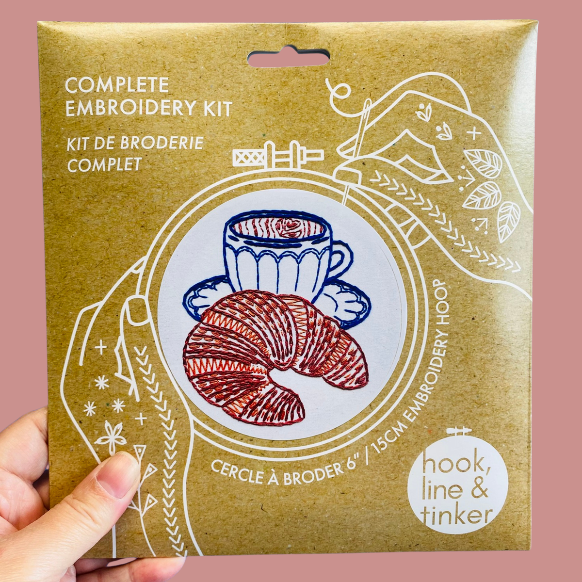 A hand holding a kraft paper package for a complete embroidery kit. The package features white line art of hands holding a teacup and croissant, with the text 'COMPLETE EMBROIDERY KIT' and 'hook, line & tinker'.