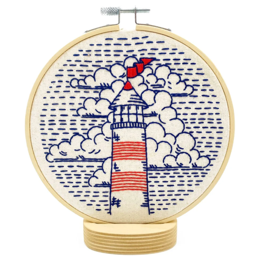 Finished embroidery project in a wooden hoop featuring a blue and red stitched design of a lighthouse against a background of clouds. 