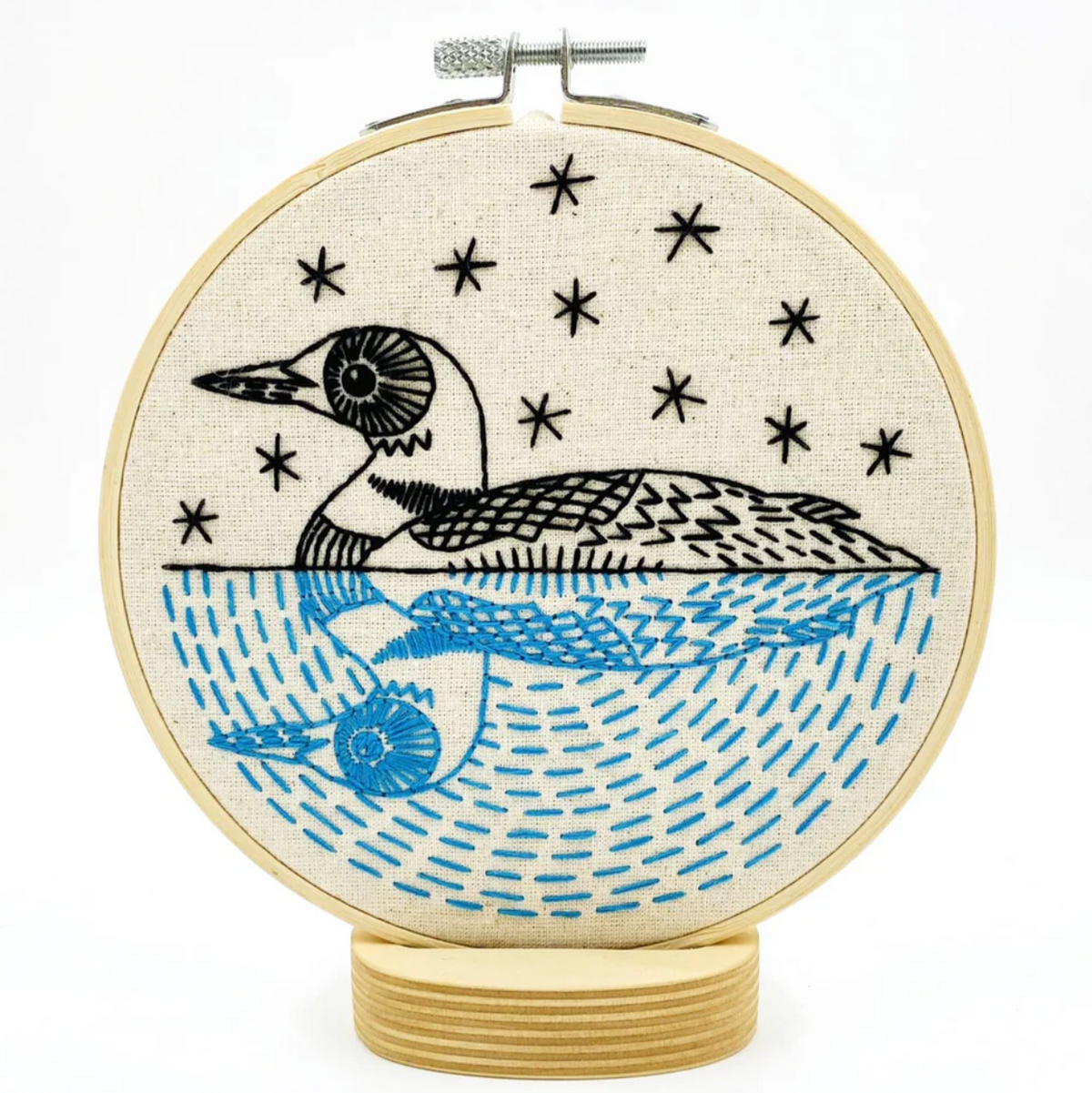 A finished embroidery project in a wooden hoop featuring a black and blue design of a loon against the stars and its reflection in the water. 