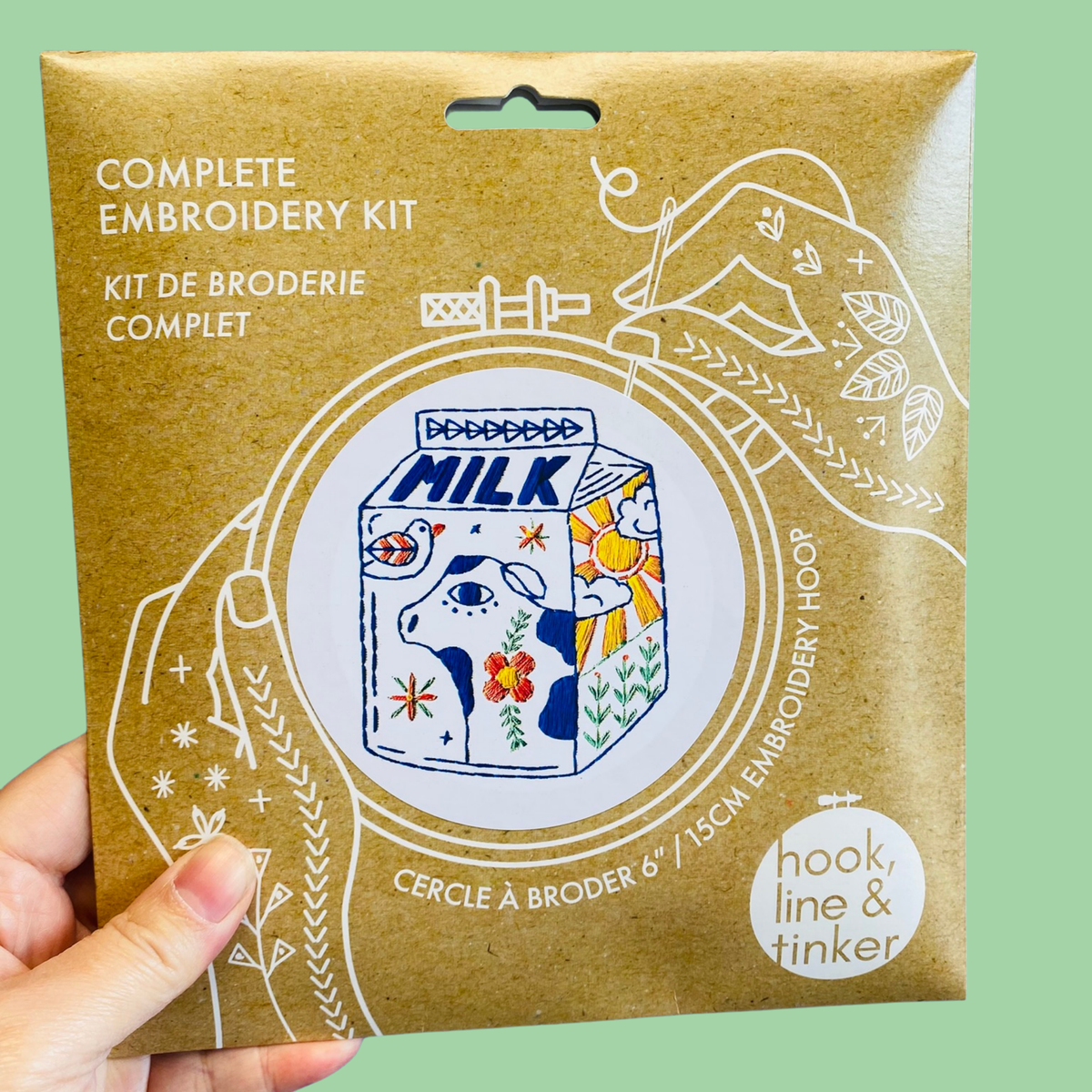 A hand holding a kraft paper package for a complete embroidery kit. The package features a colorful illustration of a milk carton with a cow and floral patterns, surrounded by white line drawings of hands and embroidery tools.