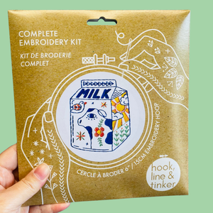 A hand holding a kraft paper package for a complete embroidery kit. The package features a colorful illustration of a milk carton with a cow and floral patterns, surrounded by white line drawings of hands and embroidery tools.