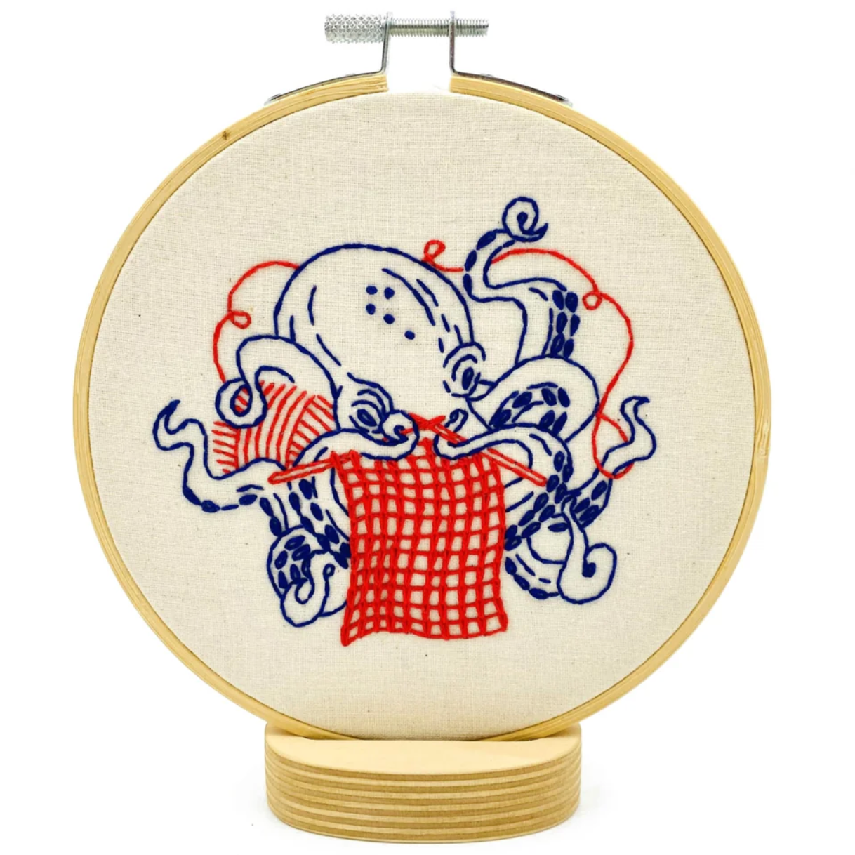 A finished embroidery project in a wooden hoop featuring a red and white design of an octopus knitting a red square. 