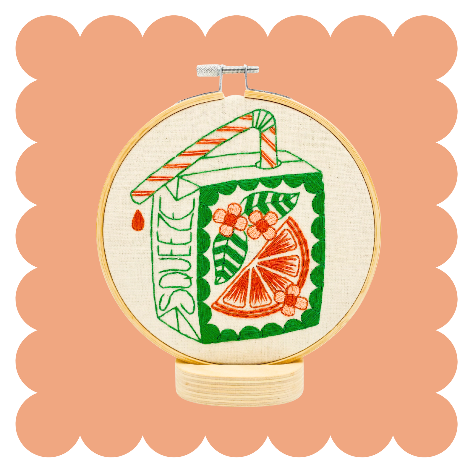A circular embroidery hoop containing a colorful stitched design of a juice carton with a striped straw and a citrus slice motif, centered on a peach scalloped background.