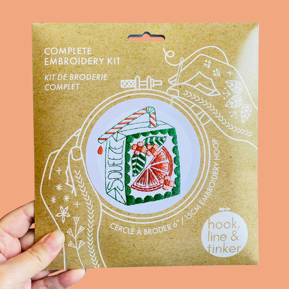 A hand holding a kraft paper package for a complete embroidery kit. The package features white line art of hands holding a hoop and an illustration of an embroidered juice box carton with green, red, and orange details.