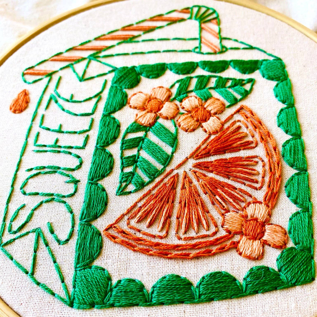 A close-up of a circular embroidery hoop showing a colorful embroidered design on light fabric. The design includes green leaves, orange flowers, a striped straw, and an orange slice motif, with the word 'SQUEEZE' stitched on the left side.
