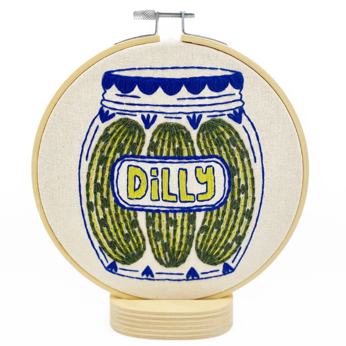 A finished embroidery project in a wooden hoop featuring a yellow, green, and blue design of a jar with three pickles in it that has a label that reads "Dilly"
