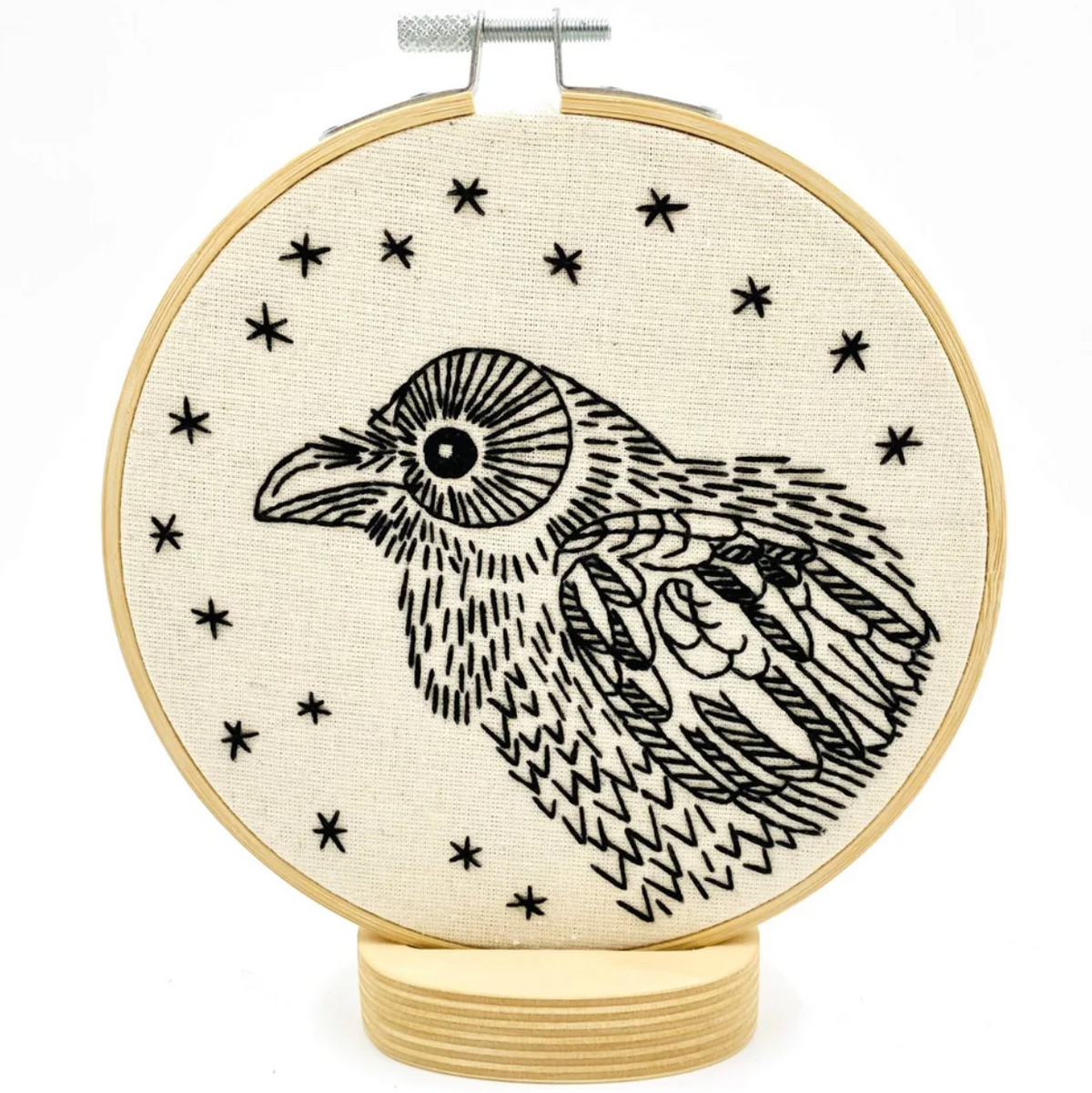A completed embroidery project in a wooden hoop, featuring a black design of a raven surrounded by stars. 