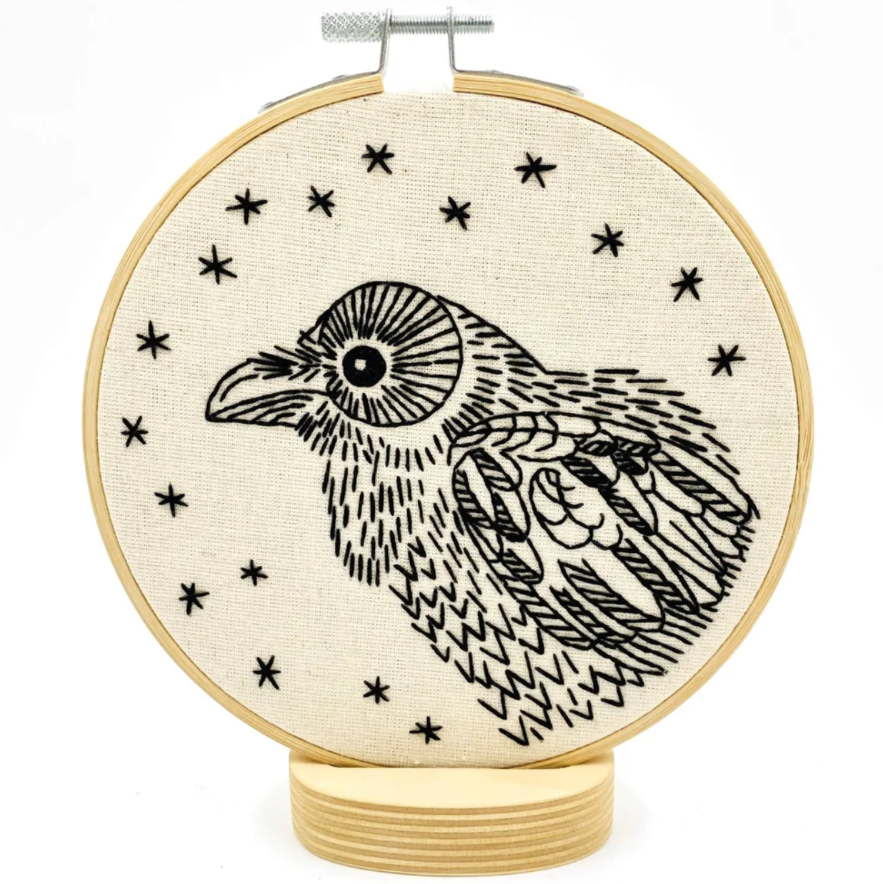 A completed embroidery project in a wooden hoop, featuring a black design of a raven surrounded by stars. 
