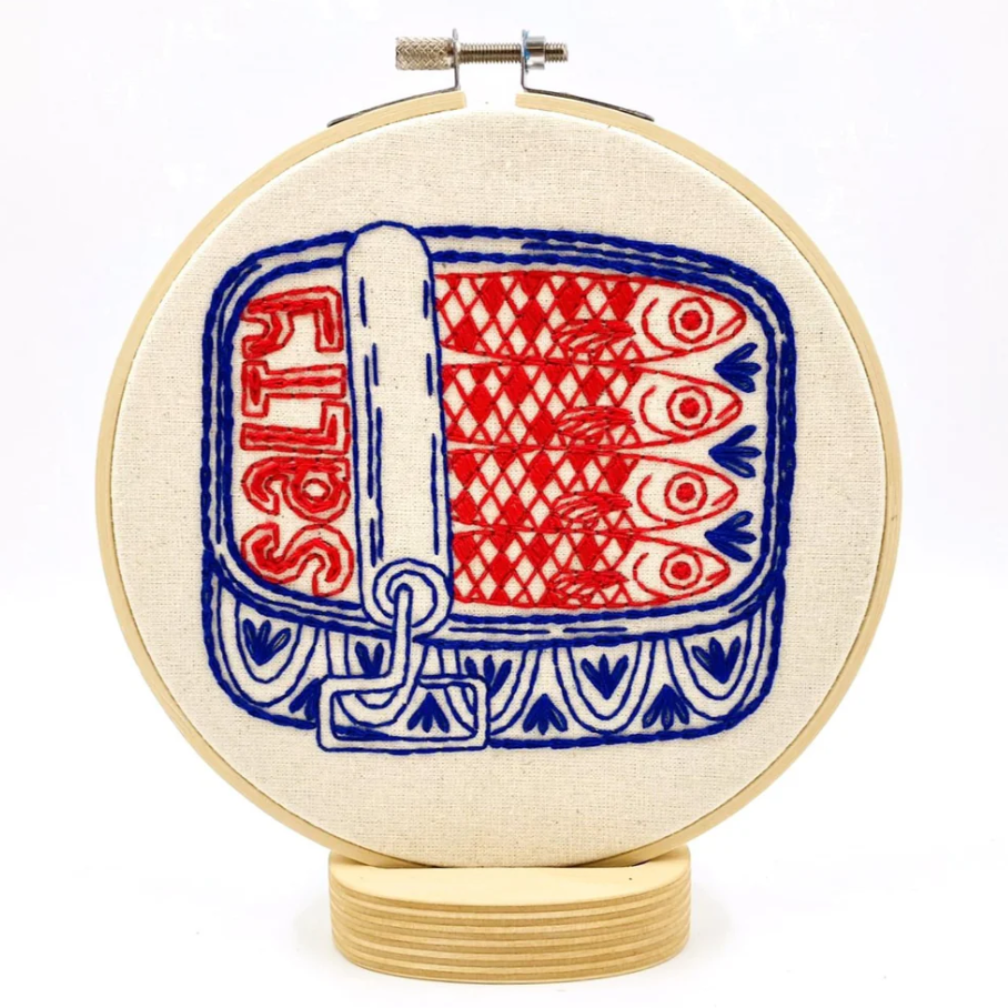 A finished embroidery project in a wooden hoop featuring a red and blue design of a tin of sardines with the text "salty" on it