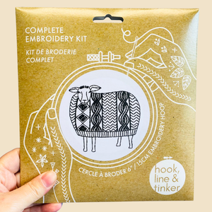 A hand holding a kraft paper package for a complete embroidery kit. The package features white line art of a patterned sheep and decorative motifs, with text reading 'COMPLETE EMBROIDERY KIT' and 'hook, line & tinker'.