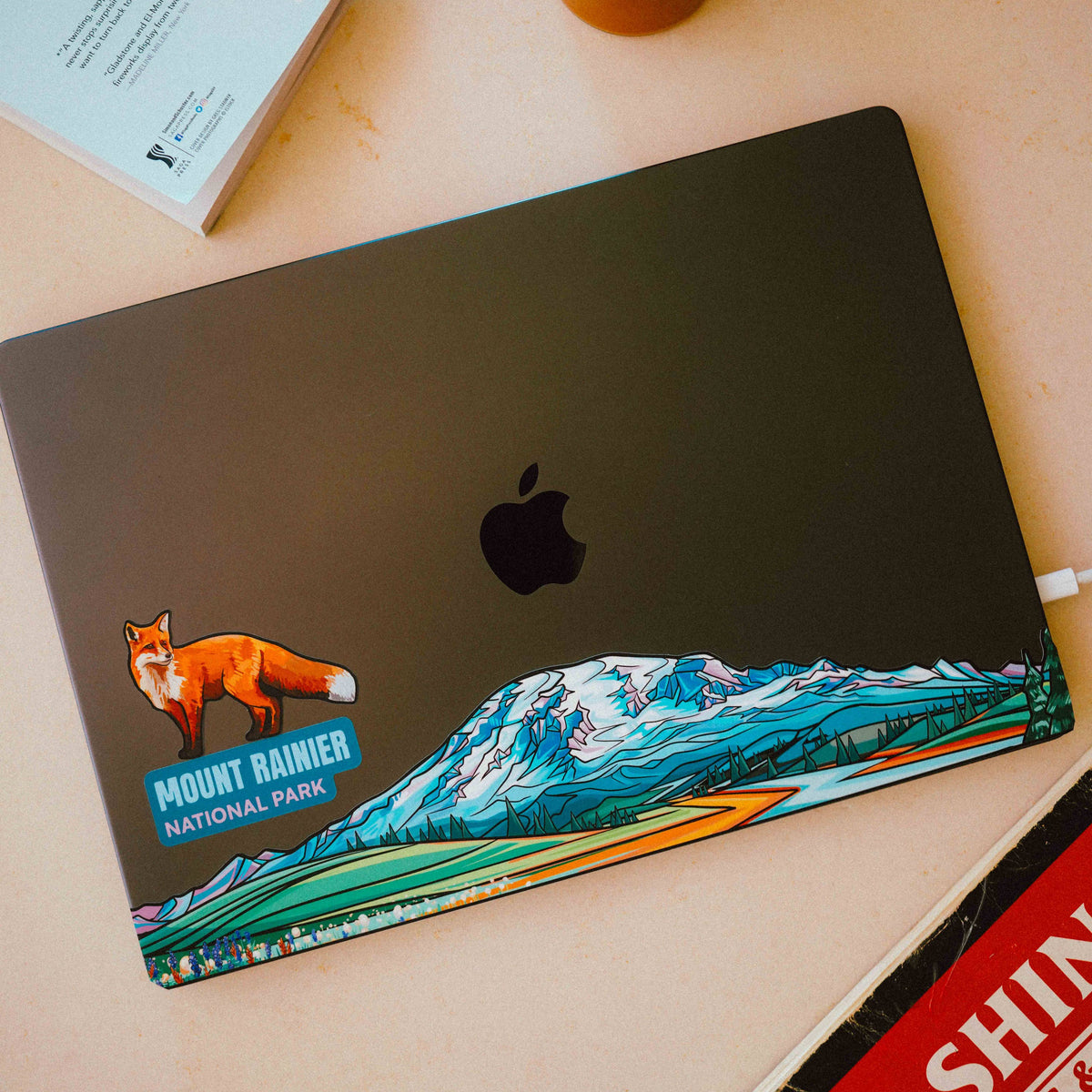 Laptop with Mount Rainier National Park sticker on a desk with a book and glass of iced coffee.