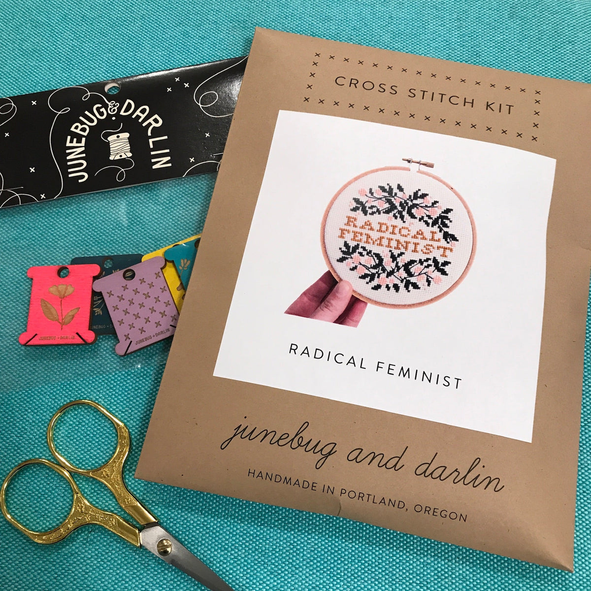 The packaging for the Radical Feminist Cross Stitch Kit by Junebug & Darlin. It is a brown kraft paper envelope with a picture of the completed kit on the front. It says Radical Feminist in the middle and is surrounded by pretty floral vines. It is sitting on a table with some embroidery scissors and a package of floss organizers. 
