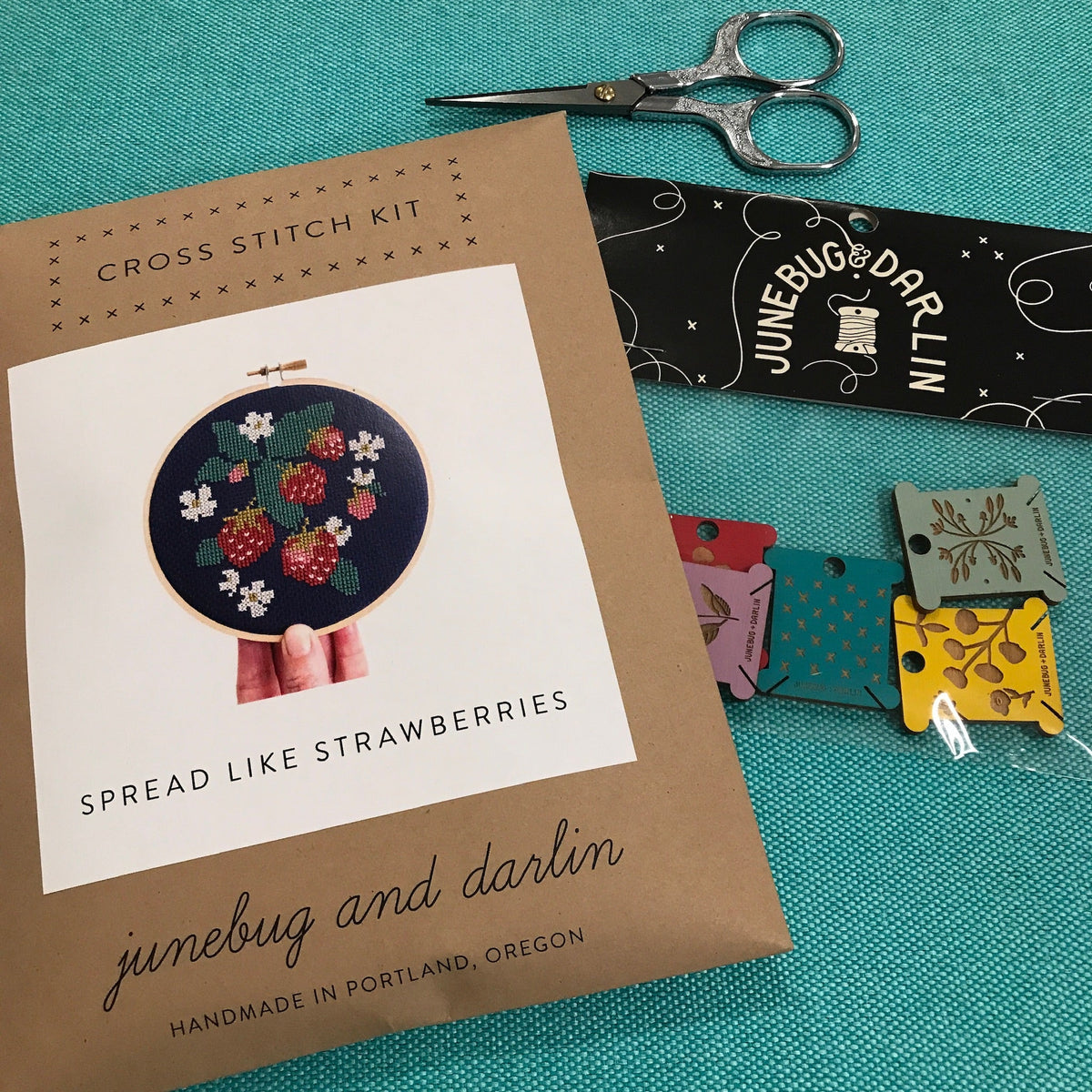 The Spread Like Strawberries kit package laid out on a table with a pair of embroidery scissors and a pack of the floss organizers. 