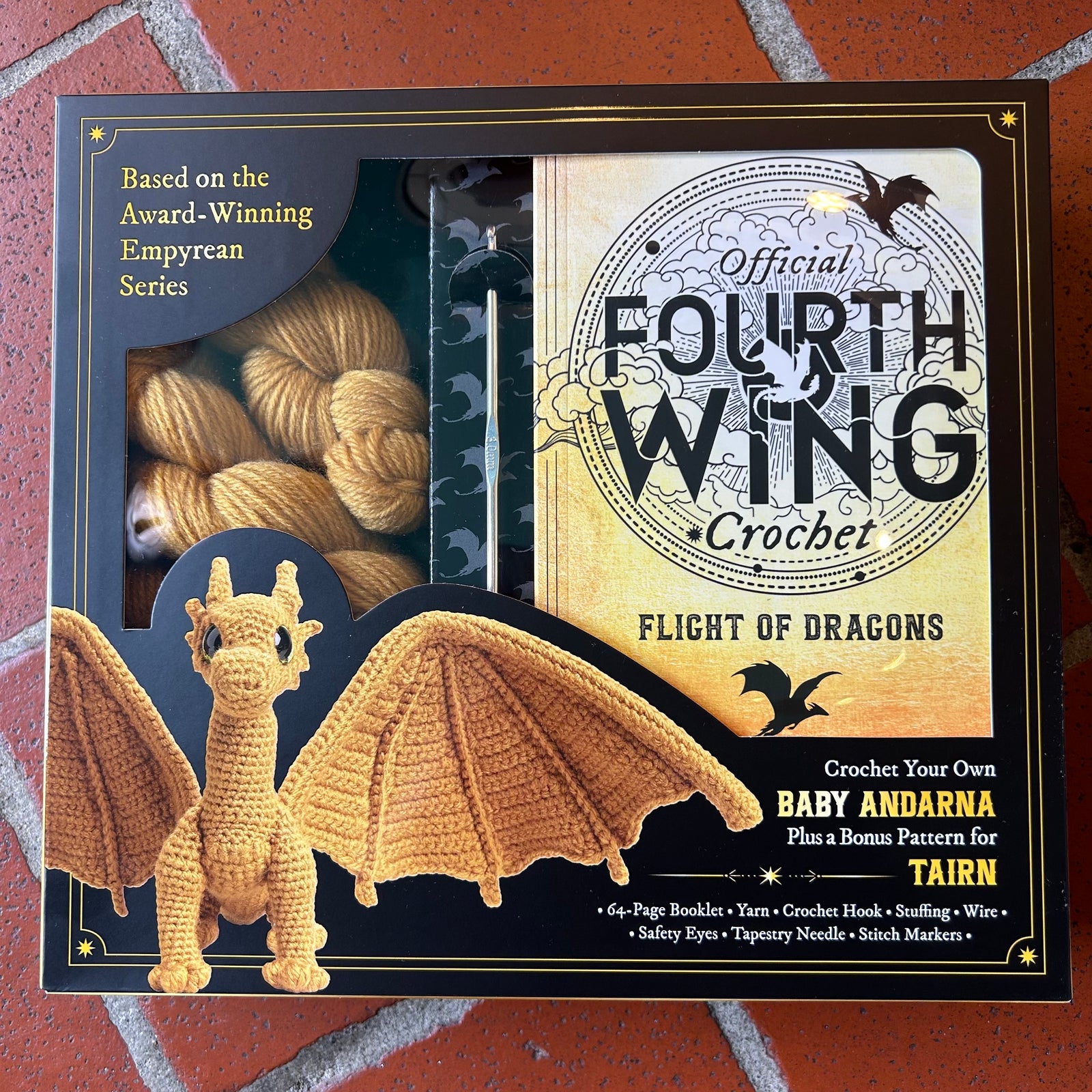 Boxed crochet kit titled 'Official Fourth Wing Crochet: Flight of Dragons' shown on a red brick surface. The package includes a crocheted dragon figure, yarn skeins in a clear window, and printed cover art with text about making a baby Andarna.